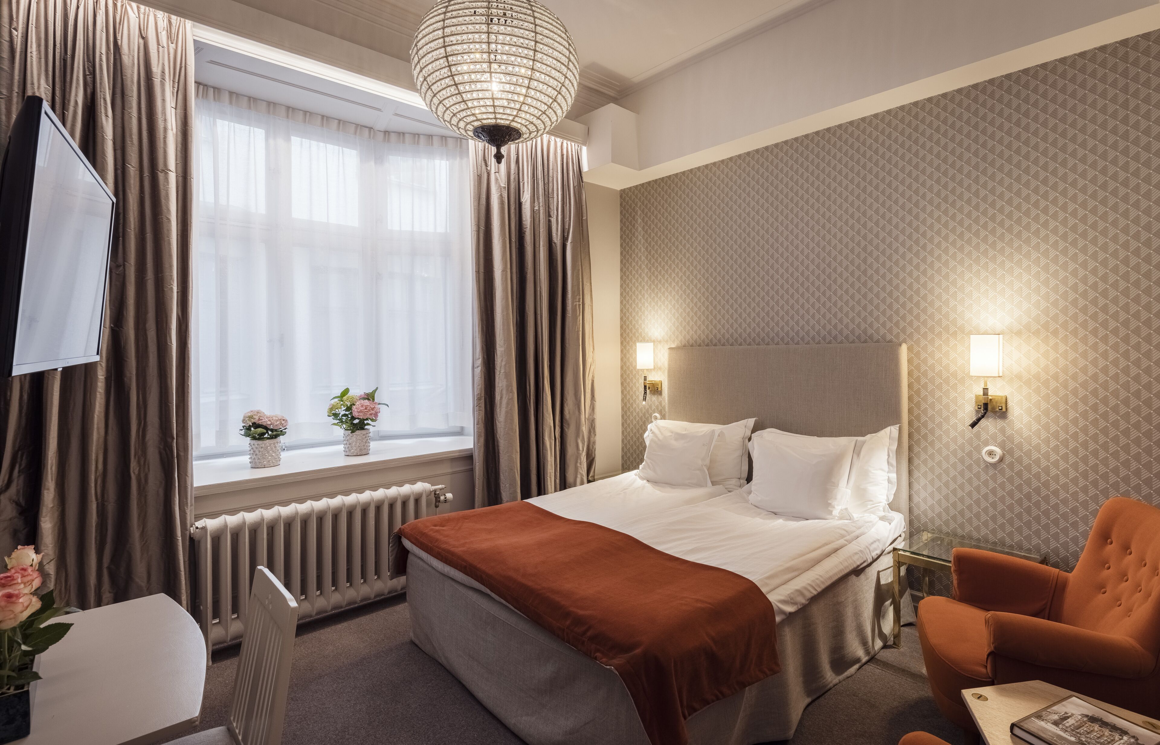 A hotel room with a bed, a chair, a table, a TV, and a window with curtains.