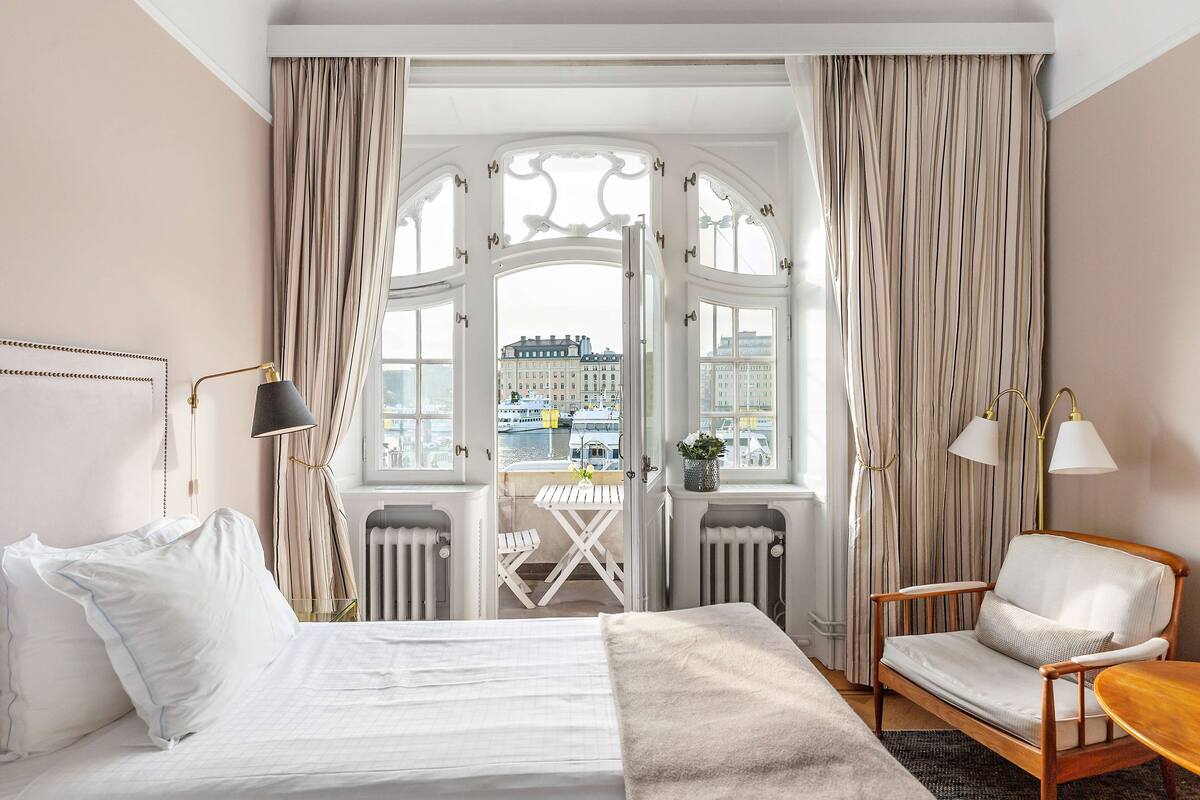 A hotel room with a bed, a chair, a small table, and a view of a cityscape through a large window.
