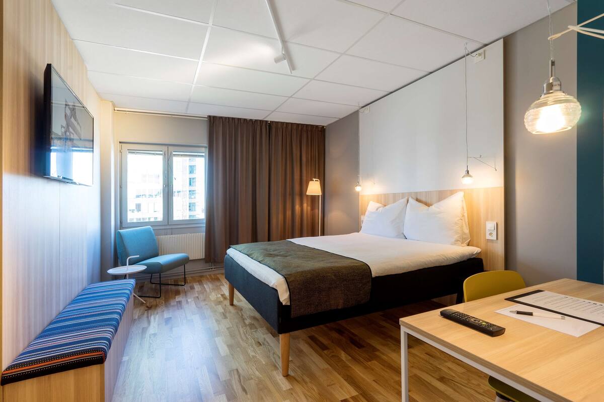 Ein Hotelzimmer mit Bett, Schreibtisch, Stuhl, Bank und Fernseher an der Wand.