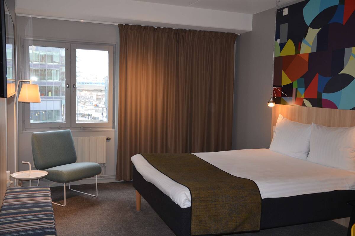 Ein Hotelzimmer mit Bett, Stuhl, kleinem Tisch und Blick auf die Stadt durch ein Fenster.