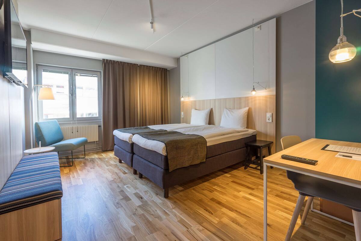 Ein modernes Hotelzimmer mit einem großen Bett, einem Schreibtisch, einem Sessel, einer Bank und einem Fenster mit Vorhängen.