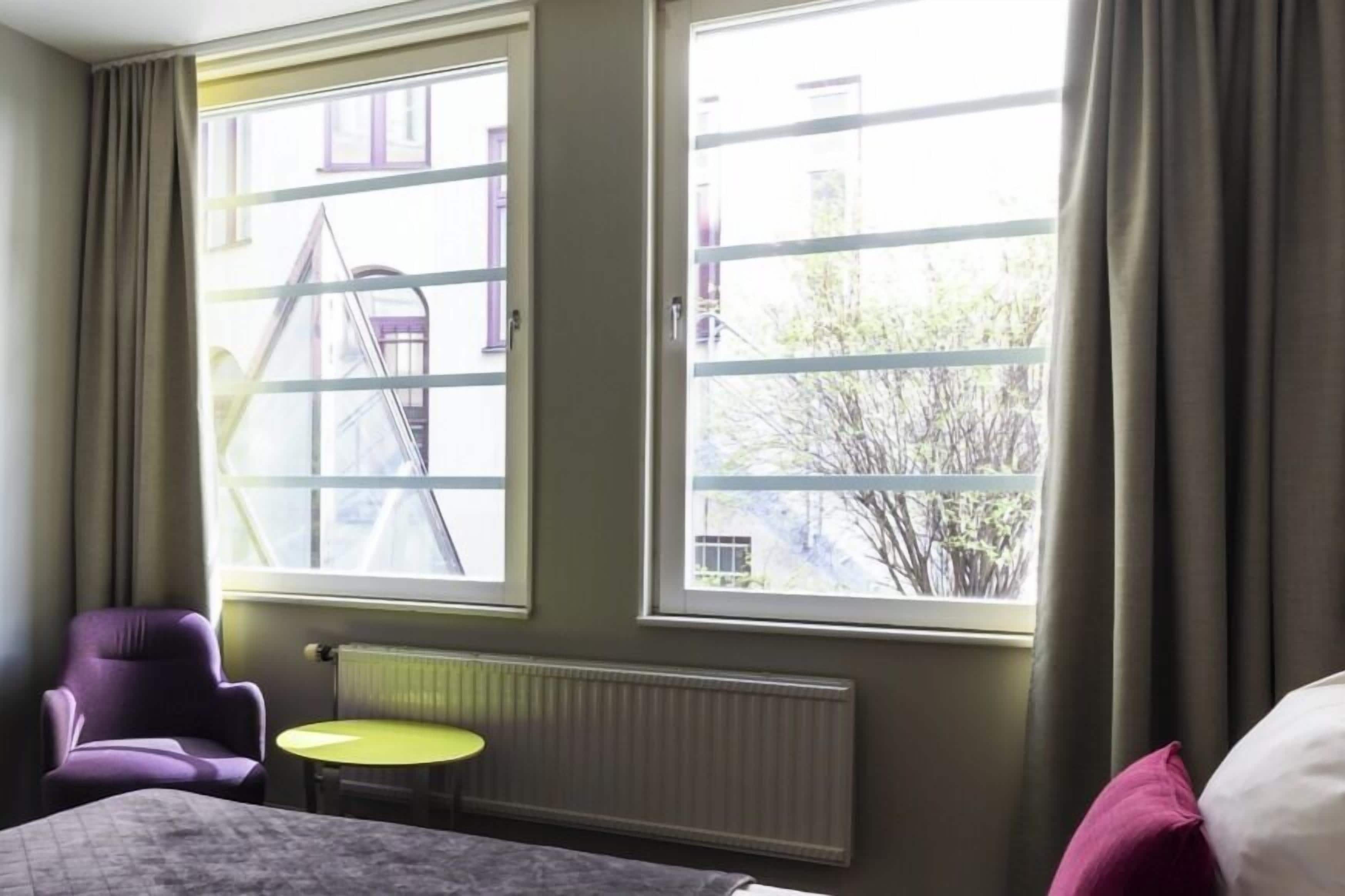 A hotel room with a bed, a purple armchair, a small yellow table, and large windows with curtains.
