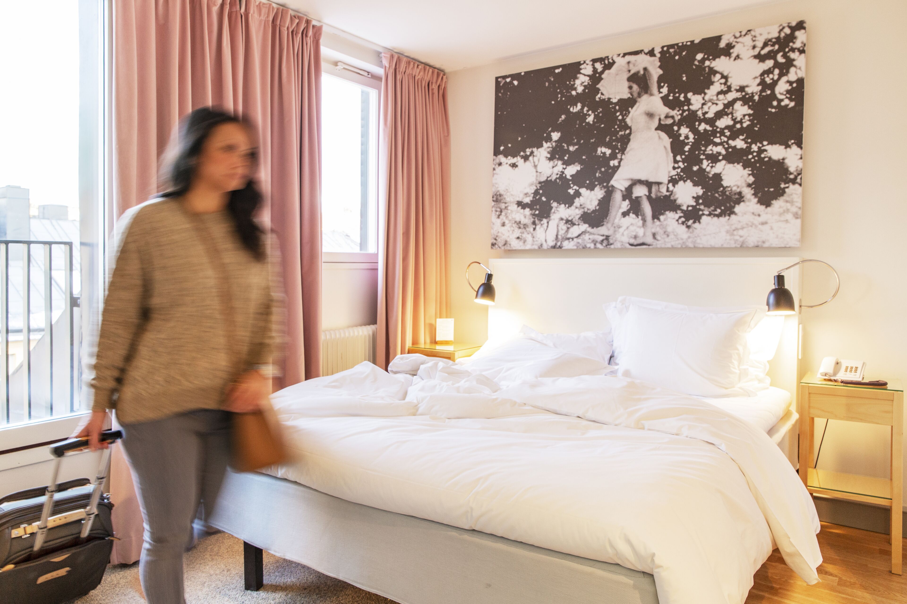 A hotel room with a bed, a nightstand with a lamp, a window with curtains, and a wall-mounted picture.