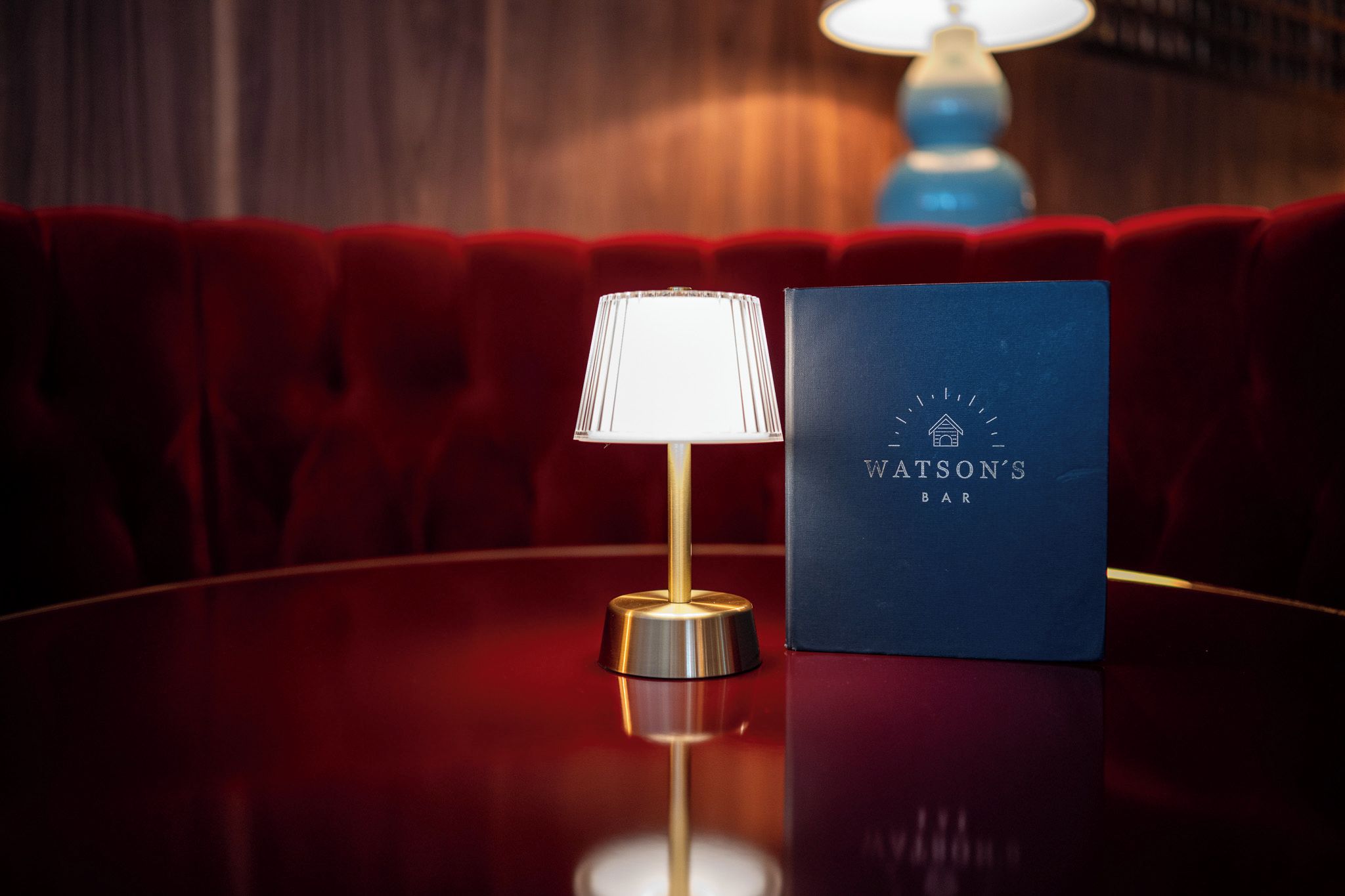 Uma mesa redonda com uma lâmpada e uma placa azul com a inscrição 'Watson's Bar' em um ambiente com assentos estofados vermelhos.