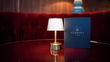 Une table ronde avec une lampe et une enseigne bleue sur laquelle on peut lire « Watson’s Bar », dans une pièce dont les sièges sont recouverts d’un tissu rouge.