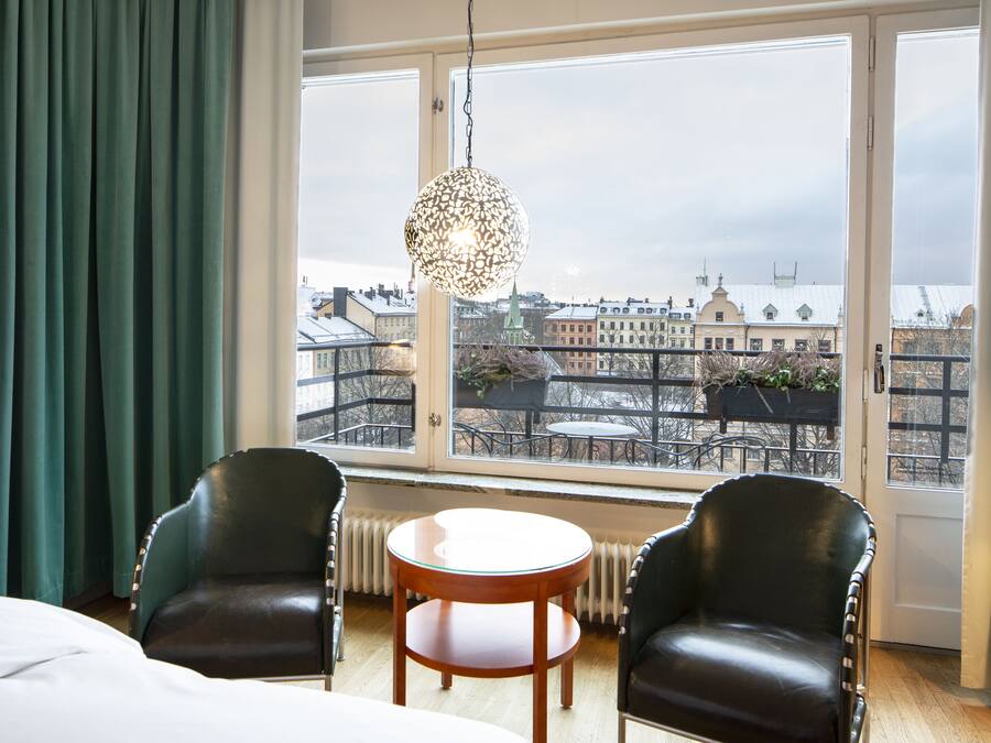A hotel room with two leather chairs, a round wooden table, and a view of a cityscape through large windows.