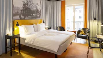 Habitación de hotel con cama, escritorio, silla, un osito de peluche y un ventanal con cortinas.