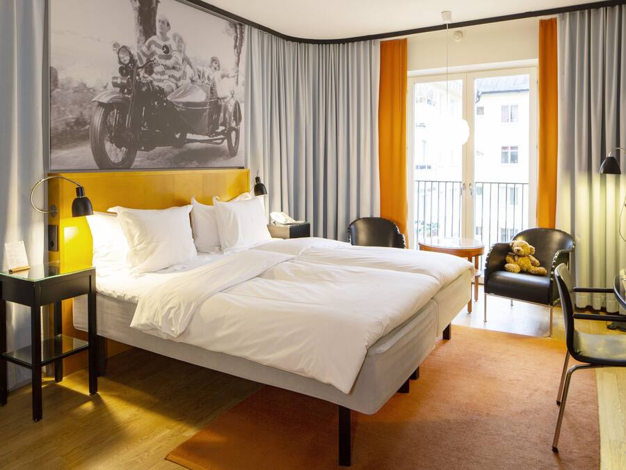 Habitación de hotel con cama, escritorio, silla, un osito de peluche y un ventanal con cortinas.