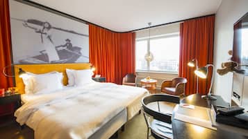 Ett hotellrum med en stor sÀng, ett skrivbord med en stol, en skrivbordslampa, ett litet bord med en lampa och ett stort fönster med röda gardiner.