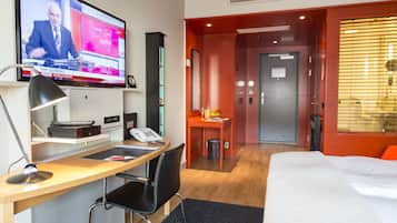 A hotel room with a desk, a bed, a TV displaying a news program, a door, and a window with blinds.