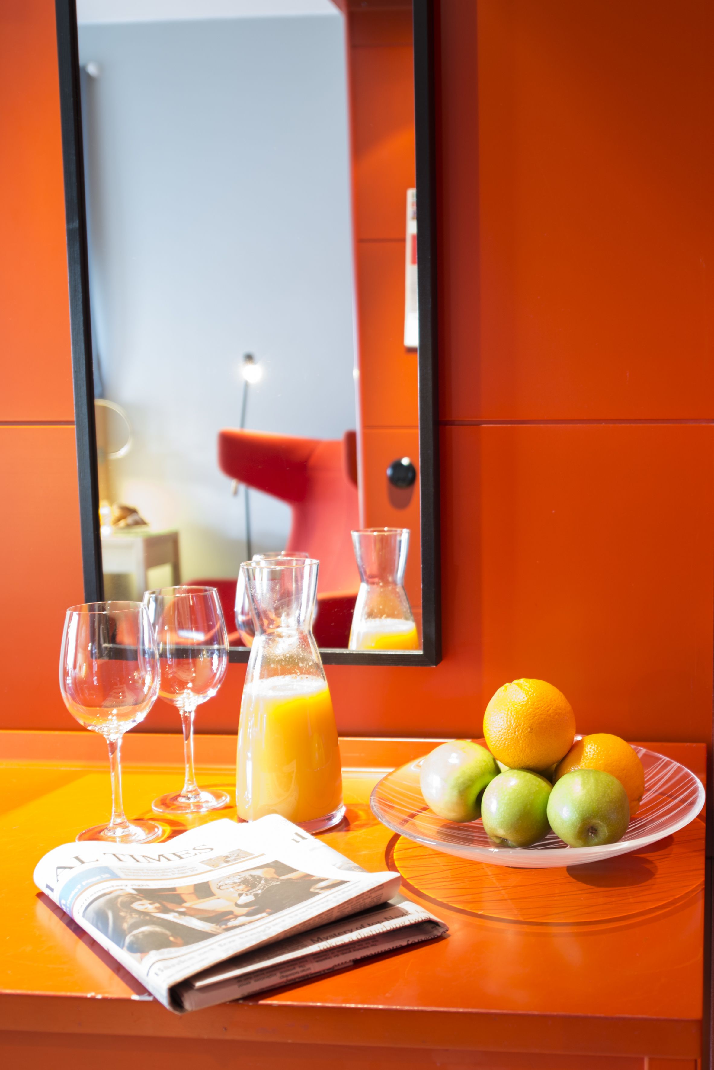 A hotel room with a table set with a newspaper, a plate of fruit, a glass of orange juice, and two empty wine glasses.