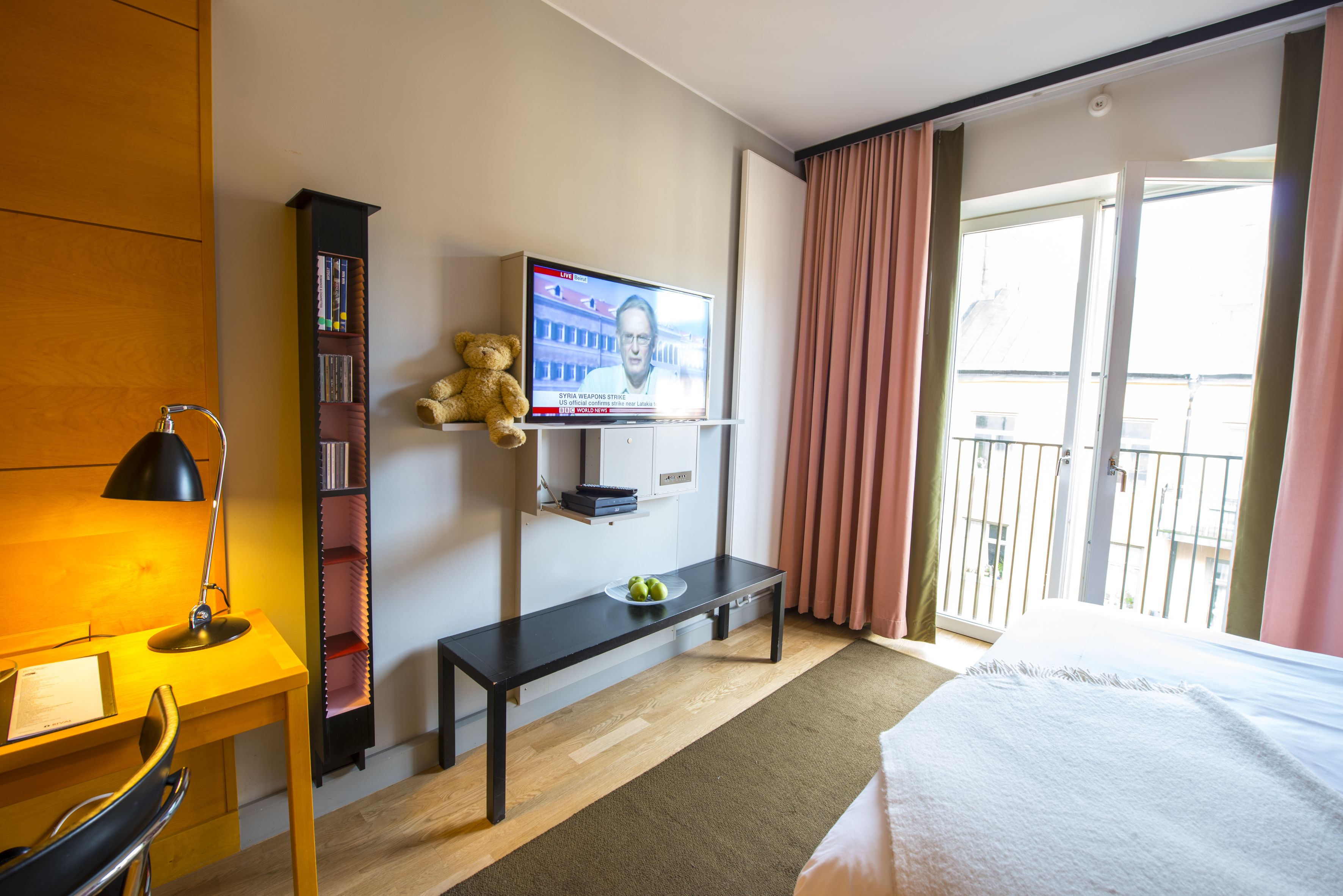 A hotel room with a bed, a desk with a lamp, a television on the wall, a bookshelf, a teddy bear, and a balcony with curtains.