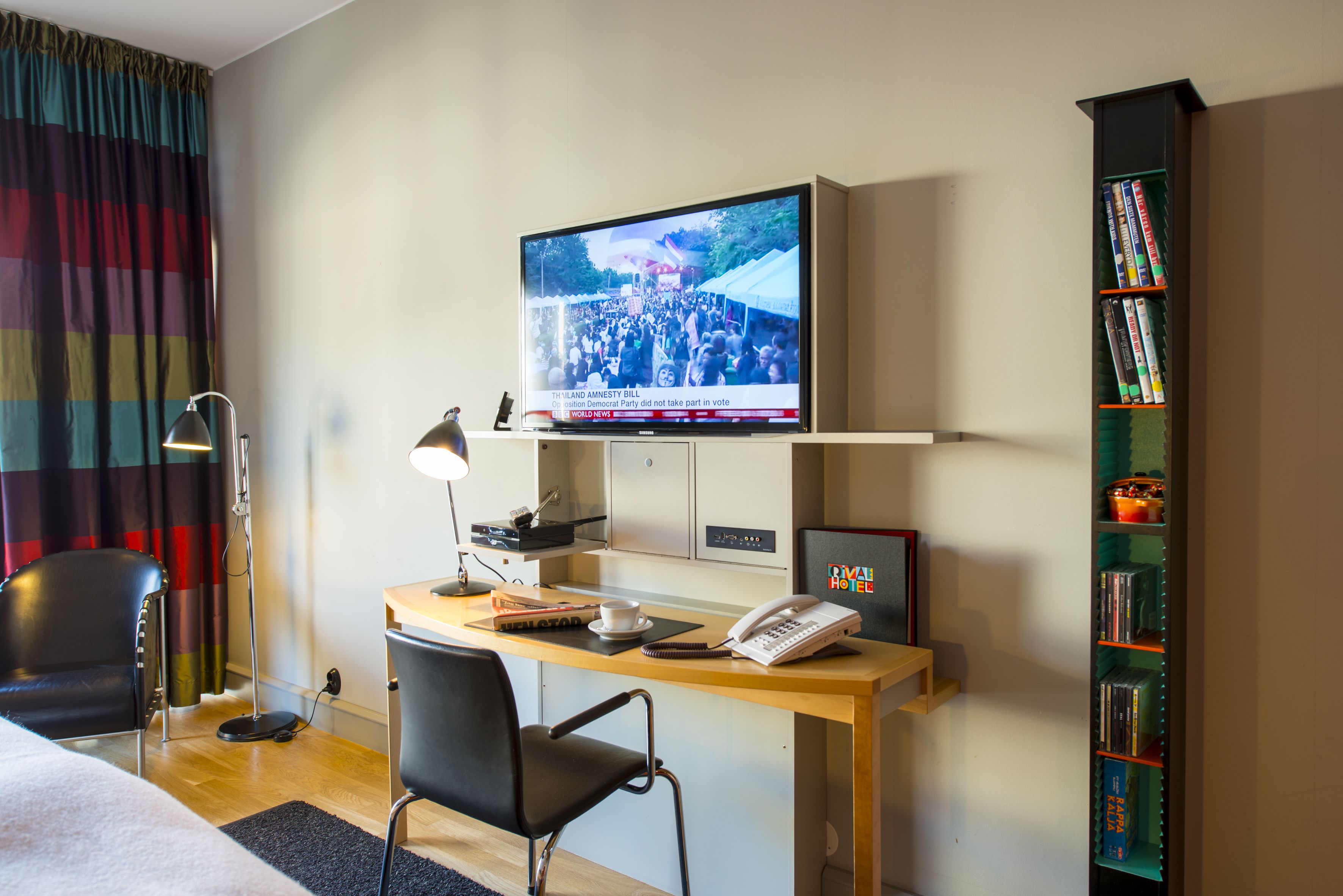 A hotel room with a desk, a television displaying a news broadcast, a bookshelf, and a bed.