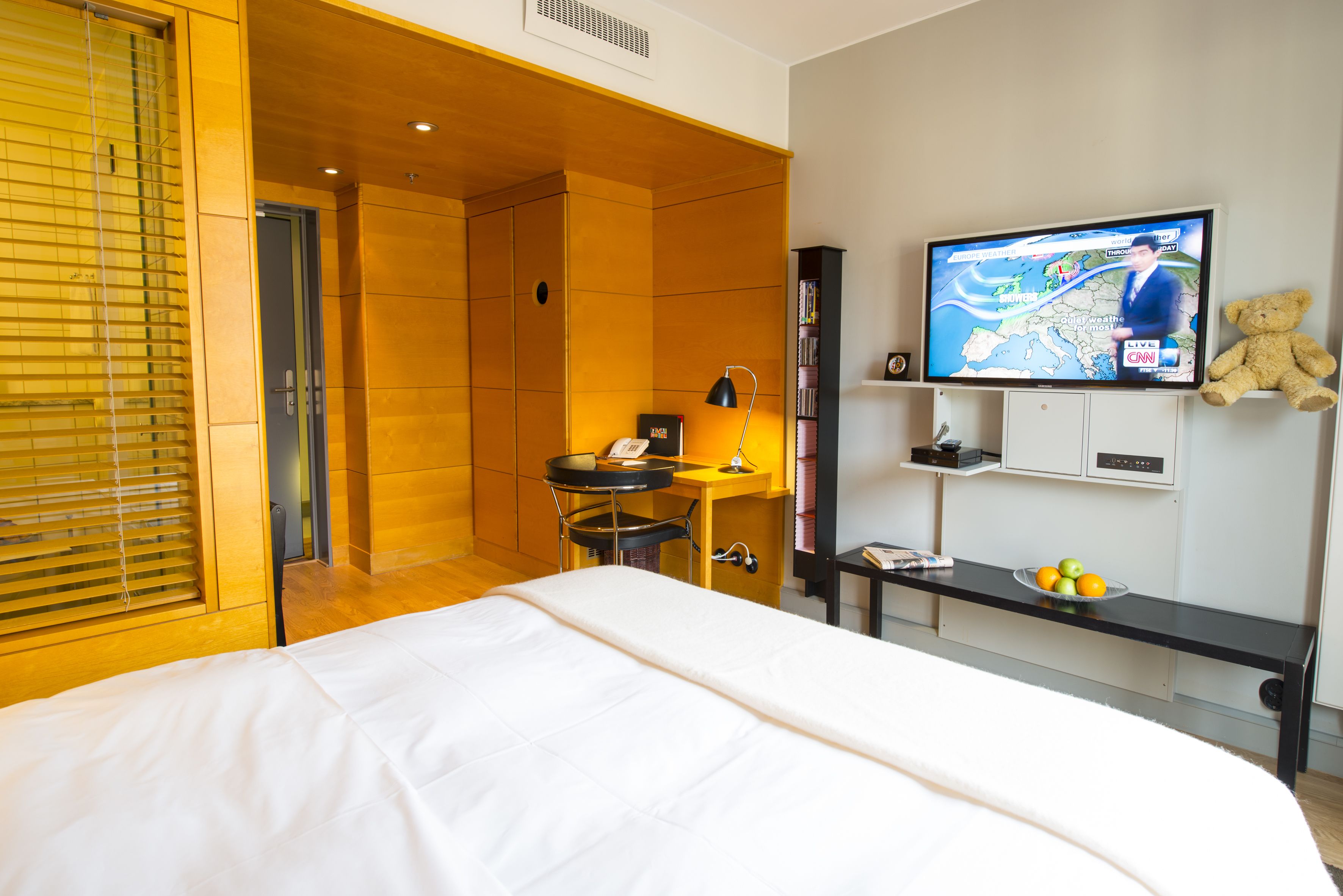 A hotel room with a bed, a desk, a television, and a teddy bear.