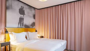 Quarto de hotel com uma cama grande, mesa de cabeceira com abajur, telefone fixado na parede e uma pintura grande acima da cama.