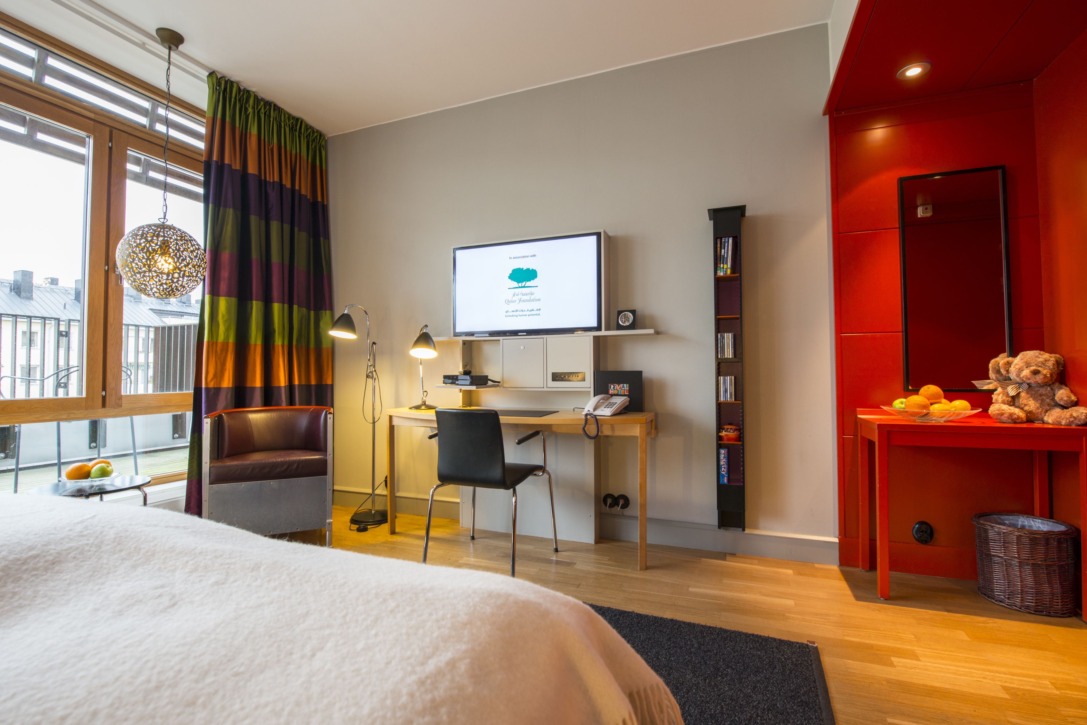 Une chambre d’hôtel moderne avec un grand lit, un bureau équipé d’un ordinateur, une chaise, une télévision et un mur d’accent de couleur rouge.