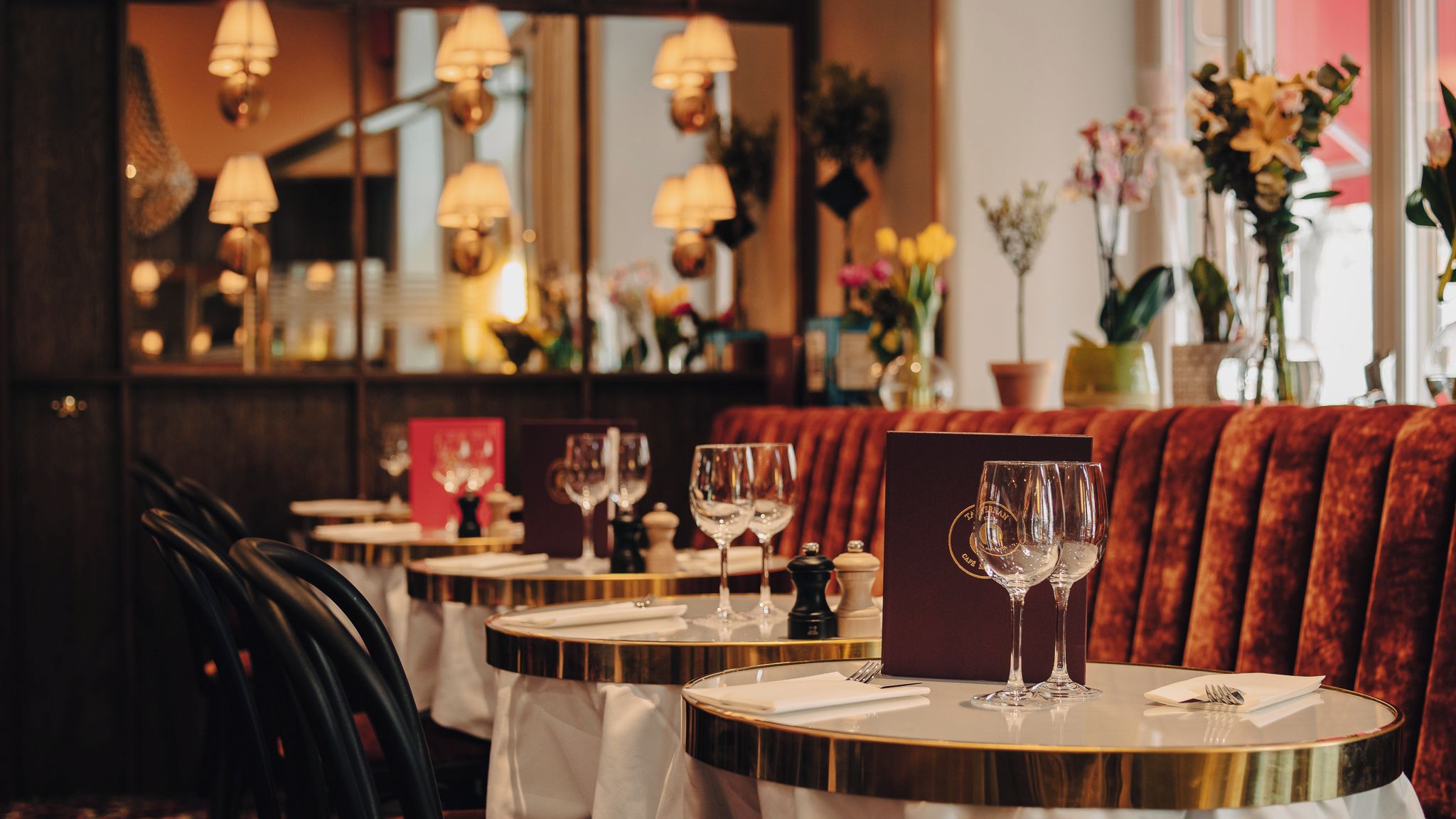 Een restaurantinterieur met ronde tafels, elk met een wijnglas en bestek, en een rode bekleden bank op de achtergrond.