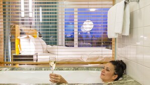 A woman in a bathtub holding a glass of champagne, with a bed and window blinds visible in the background. - Hotel Rival (Stockholm)
