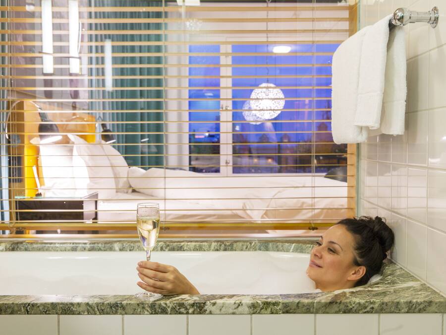 Una mujer en una bañera sosteniendo una copa de champán, con una cama y persianas visibles al fondo.