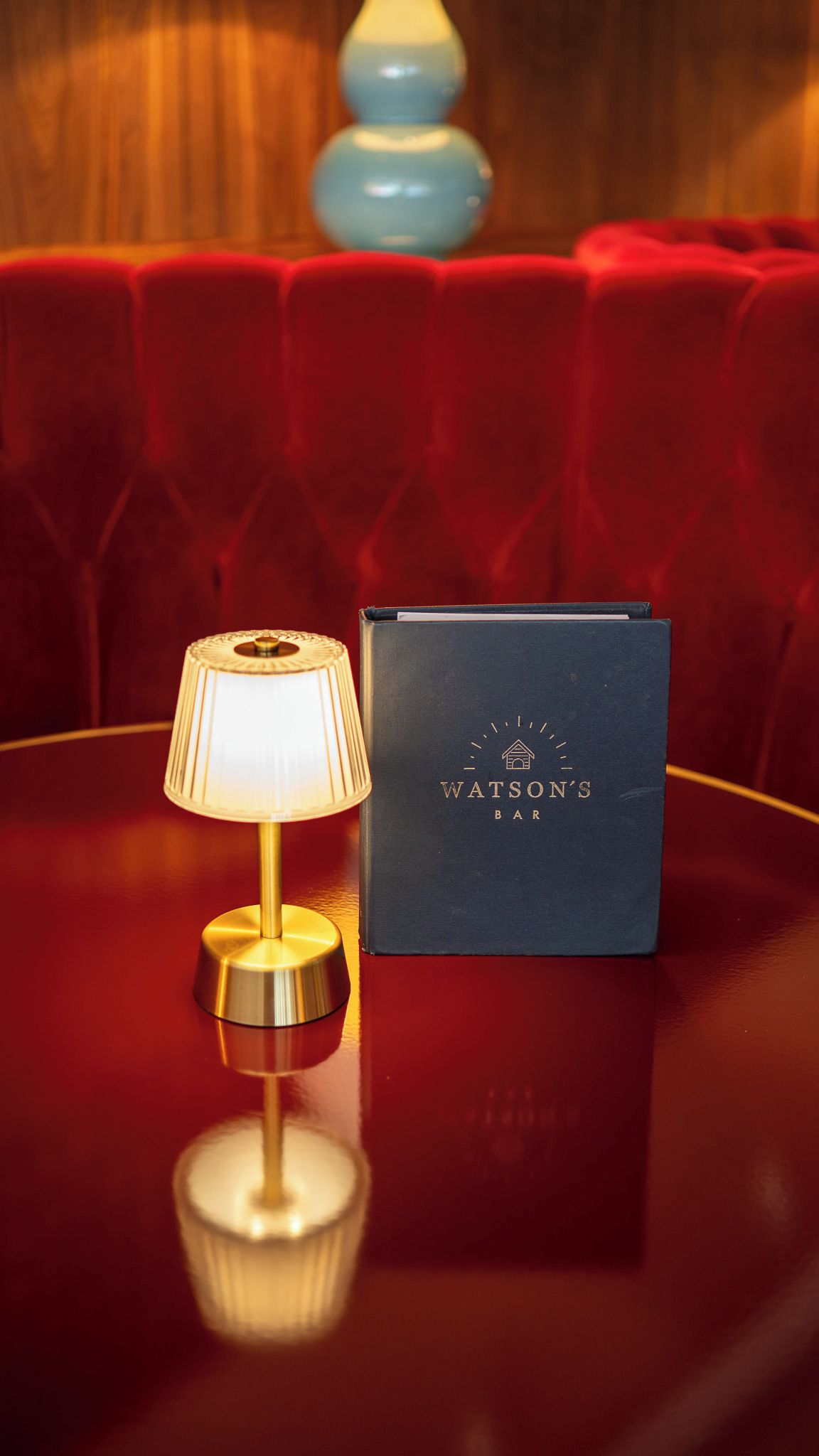 Un canapé en velours rouge, avec une lampe et un livre intitulé « Watson’s Bar », posés sur une surface réfléchissante.