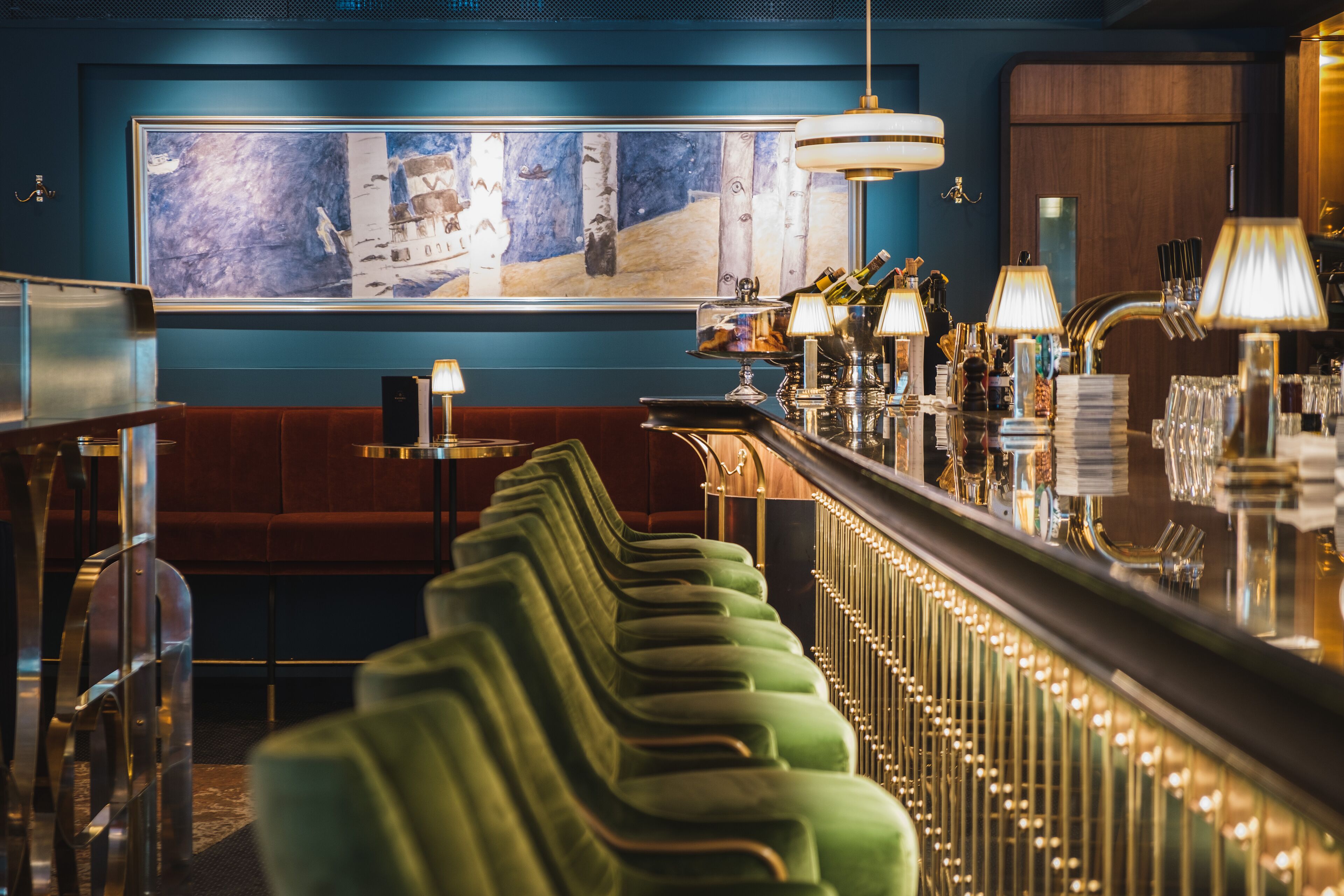 Un espace bar doté de sièges rembourrés verts, d’un grand tableau abstrait accroché au mur et d’un comptoir de bar bien éclairé, où sont exposées diverses bouteilles et différents types de verres.