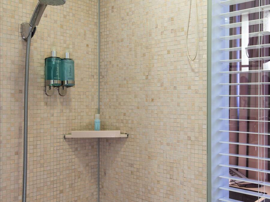 Zona de ducha con azulejos, ducha de mano, repisa de vidrio y ventana con persianas.