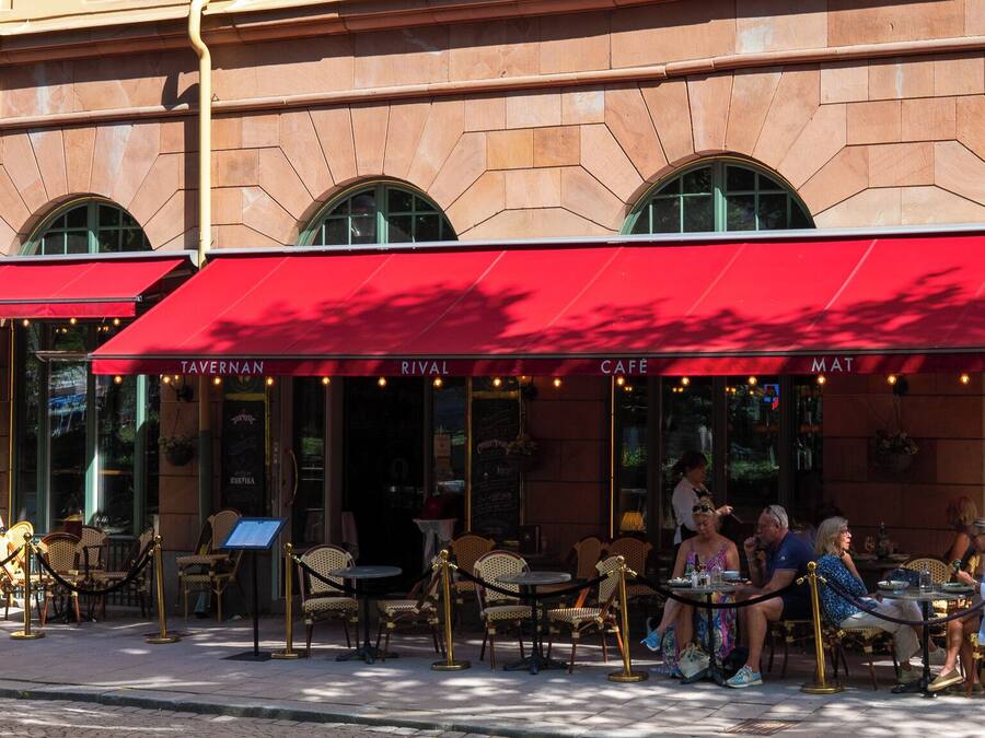 Un café al aire libre con toldos rojos, mesas en la terraza y fachada de edificio de piedra.