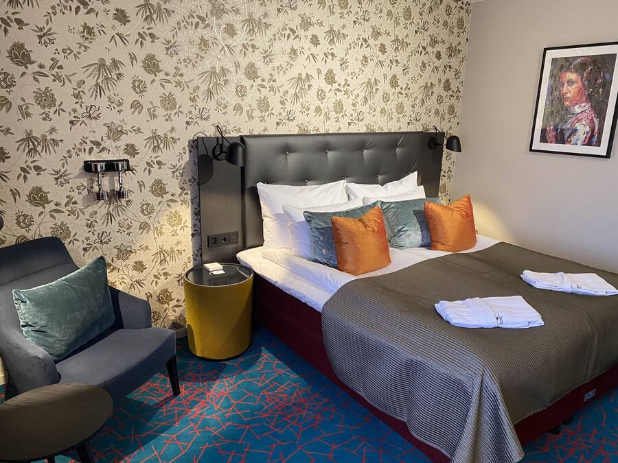 A hotel room with a tufted headboard, a bed with white and multicolored pillows, a gray sofa, a yellow side table, and a patterned carpet.