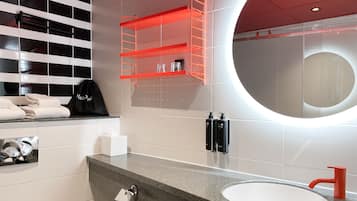 A modern bathroom with a granite countertop, a round mirror, a red towel rack, and a black soap dispenser.