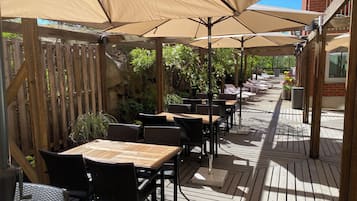 An outdoor patio with wooden tables and chairs, shaded by large umbrellas, and a wooden fence enclosing the area.