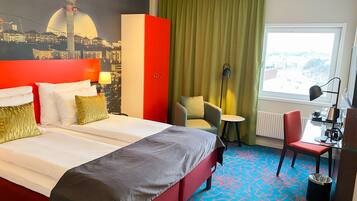 A hotel room with a bed, a desk, a chair, a lamp, a radiator, and a view of the cityscape.