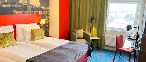 Une chambre d’hôtel avec un lit, un bureau, une chaise, une lampe, un radiateur et une vue sur le paysage urbain.