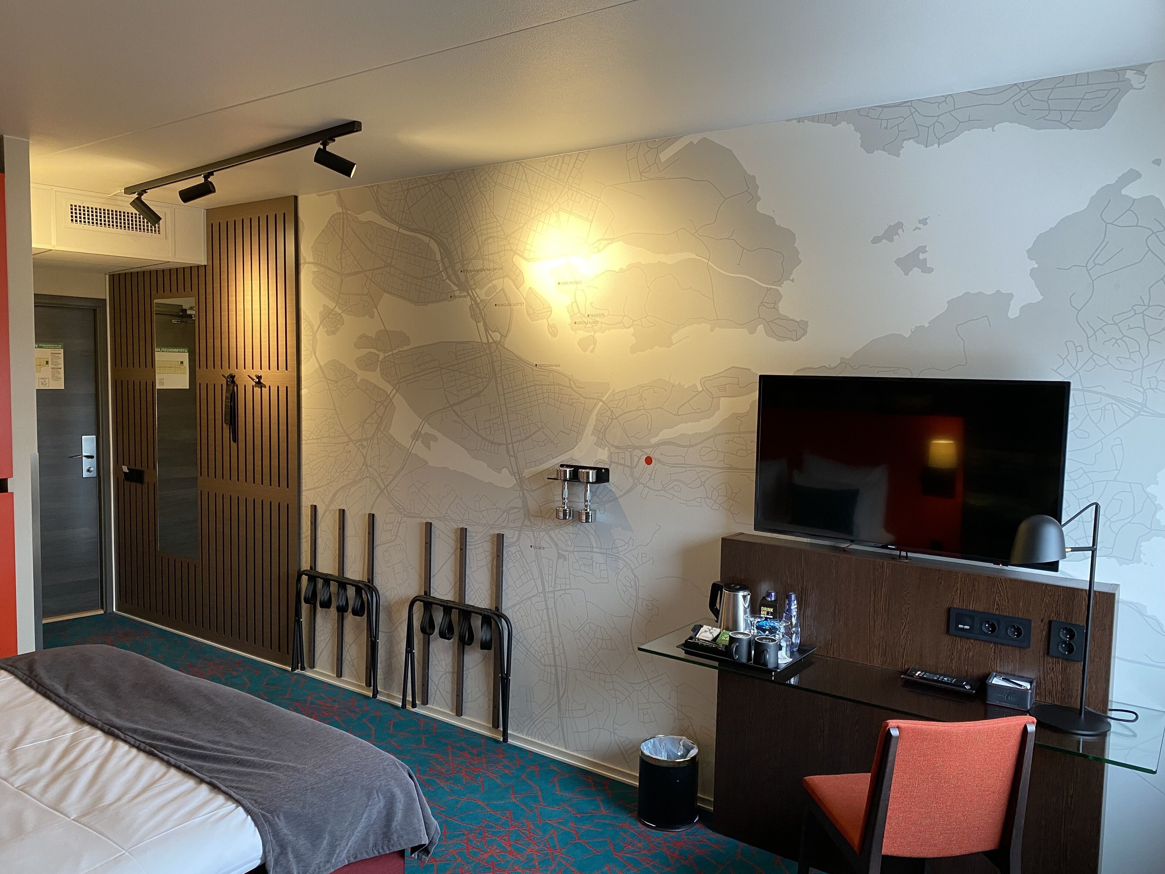 A hotel room with a bed, a desk with a TV, a map on the wall, and a coat rack.