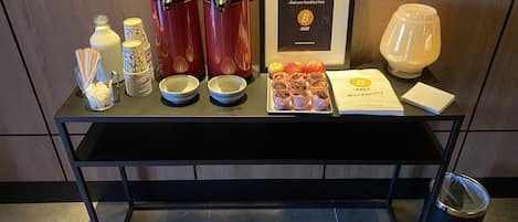 A breakfast buffet setup with coffee makers, bowls, apples, muffins, and a framed menu.