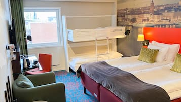 Habitación de hotel con literas, un escritorio con silla, un sofá rojo y un ventanal con vista a un paisaje urbano.