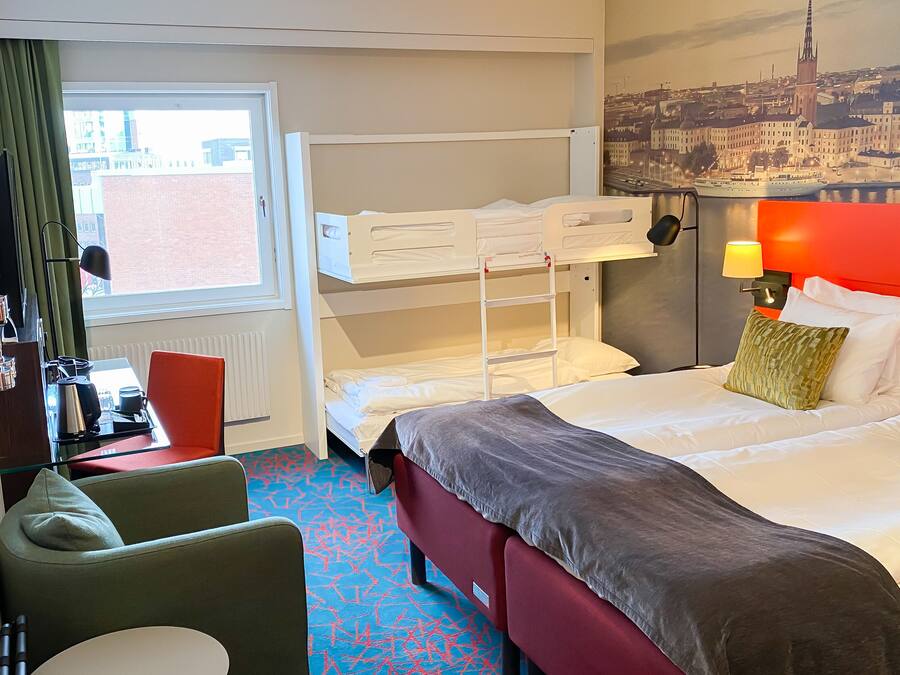 Habitación de hotel con literas, un escritorio con silla, un sofá rojo y un ventanal con vista a un paisaje urbano.