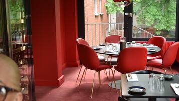 Comedor con sillas y mesas rojas, un expositor de vidrio y vista a un edificio de ladrillo y árboles.