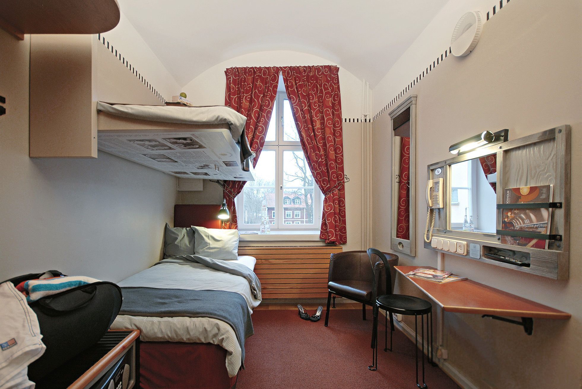 Ett kompakt hotellrum med en våningssäng, ett skrivbord med en stol, ett litet bord och en spegel.