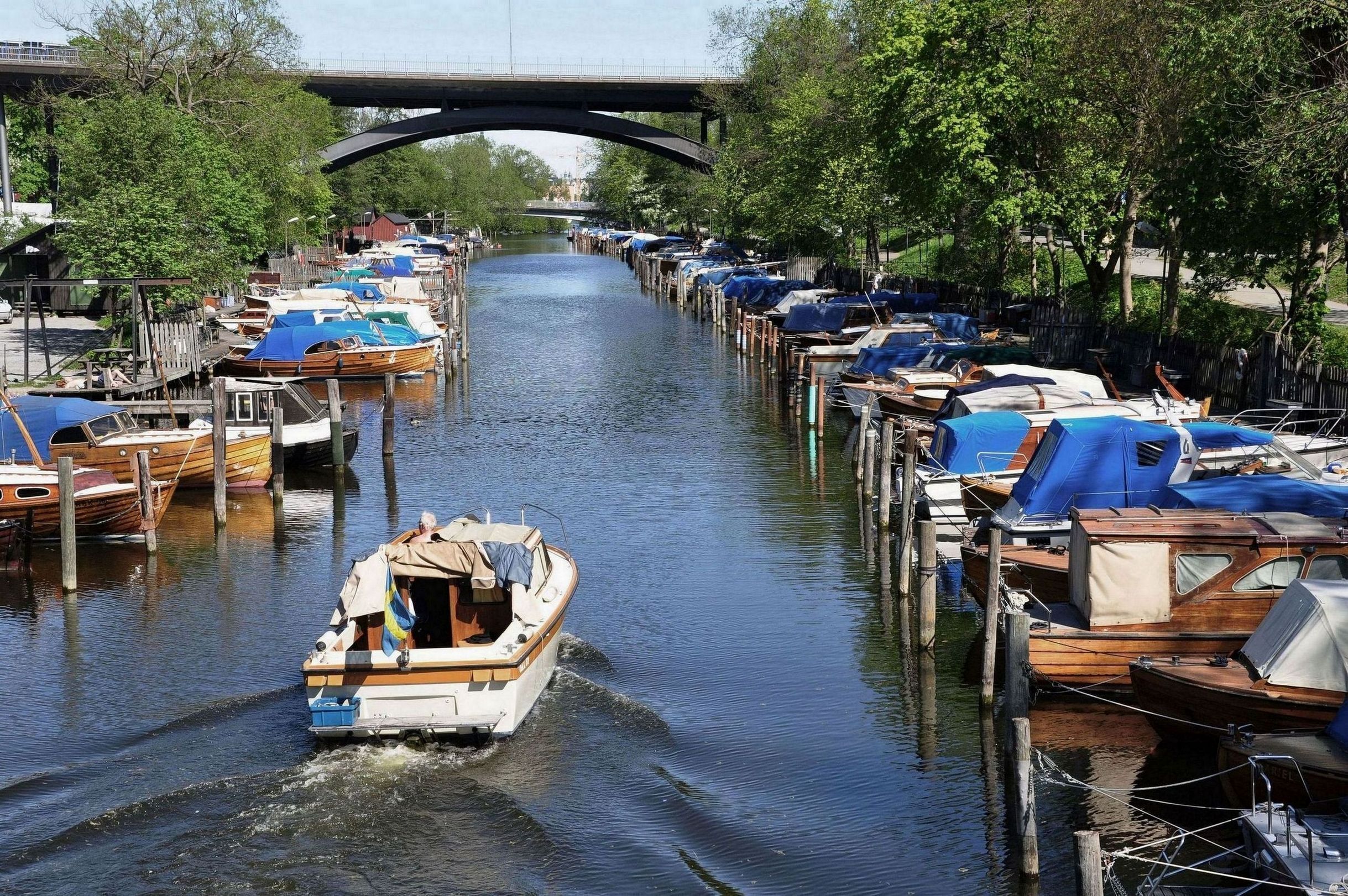 En kanal med båtar förtöjda längs båda sidorna, en bro i bakgrunden och en båt som färdas på vattnet.