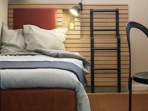 Una cama con cabecera, almohadas, una mesita de noche con lámpara y una silla.