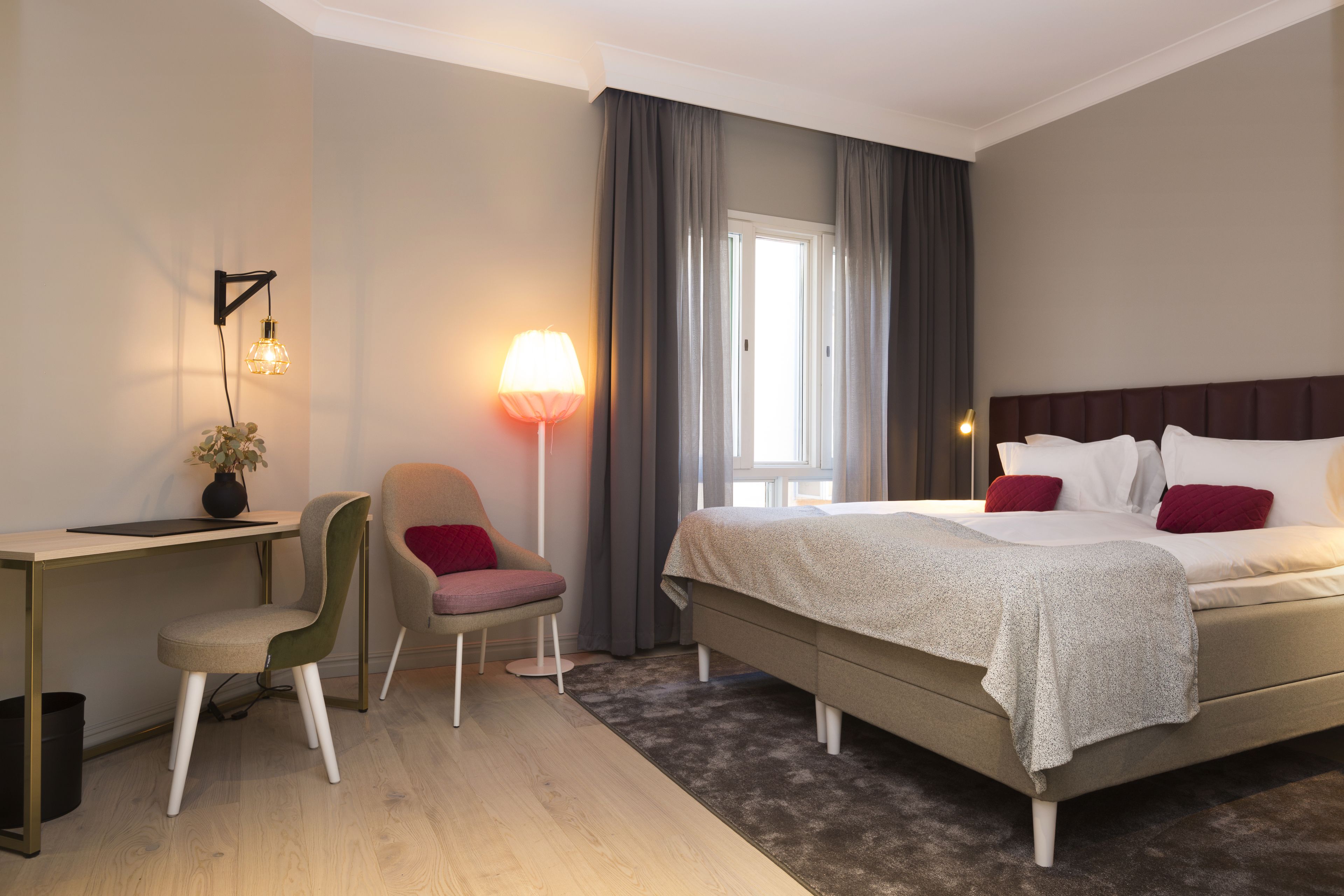 A hotel room with a bed, a desk, a chair, a lamp, and a window with curtains.