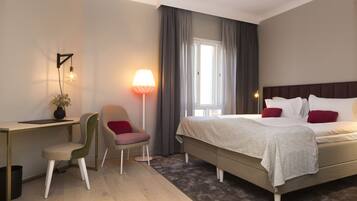 A hotel room with a bed, a desk, a chair, a lamp, and a window with curtains.