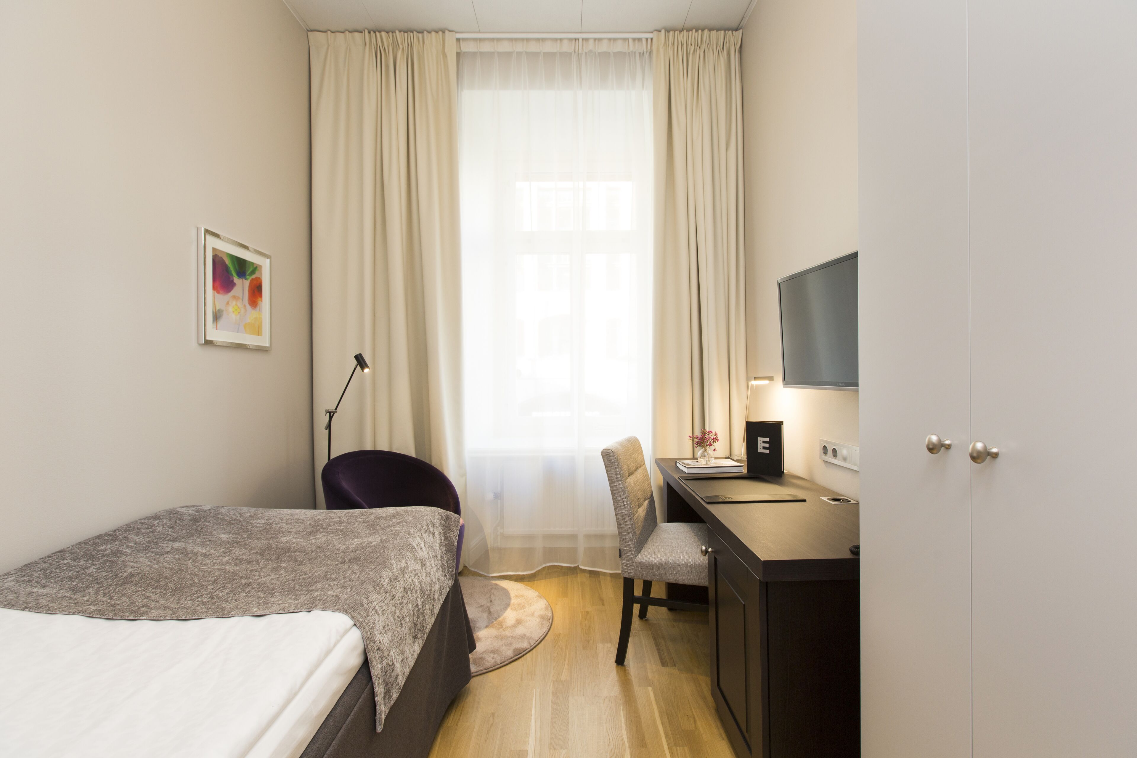A hotel room with a bed, a desk, a chair, a lamp, a TV, and a window with curtains.