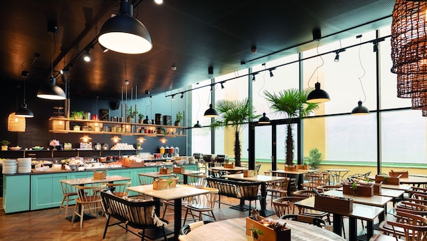 Un restaurante con mesas y sillas de madera, una zona de bar con estantes llenos de artículos y ventanas amplias que dejan entrar luz natural.