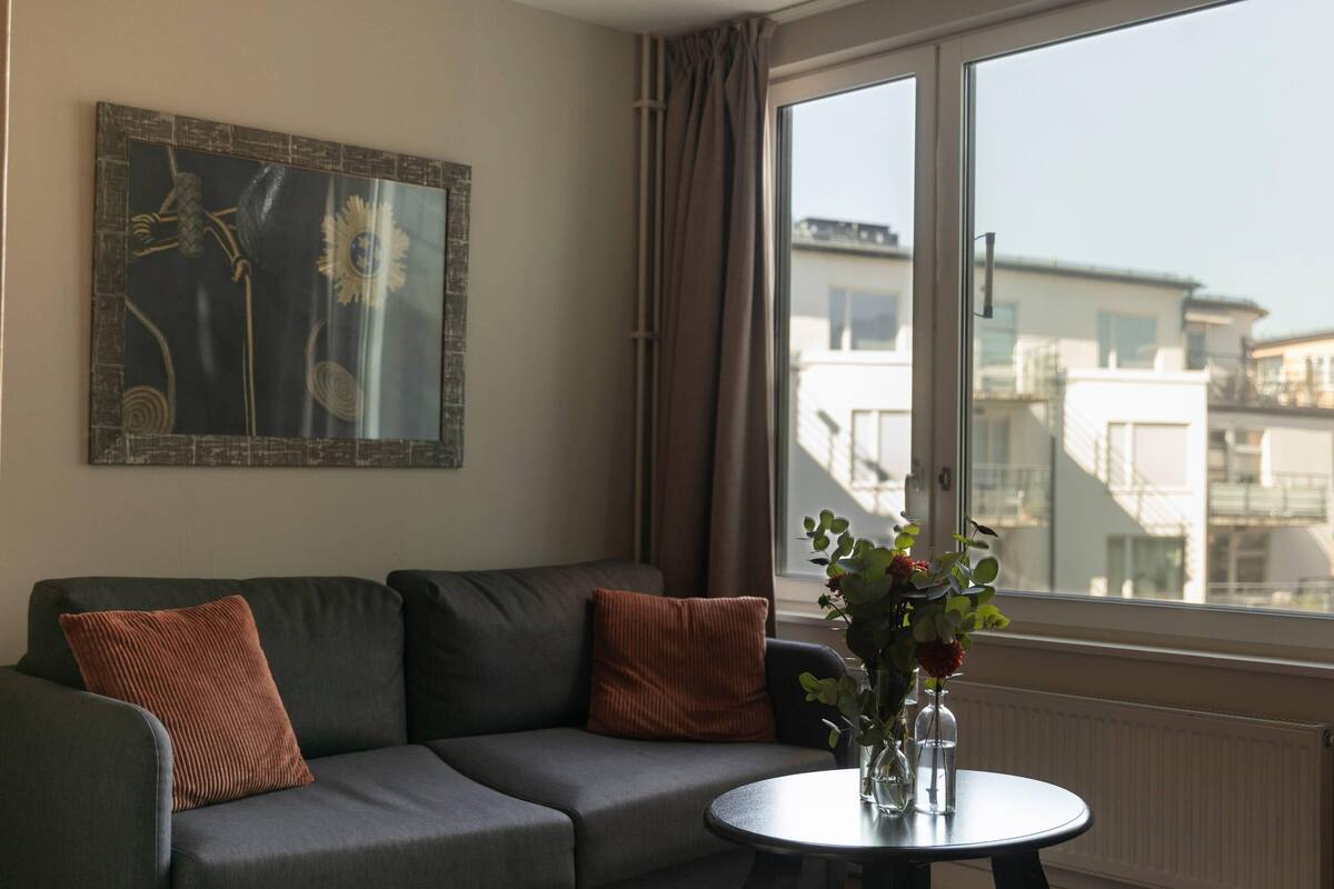Ein modernes Wohnzimmer mit einem grauen Sofa, zwei braunen Kissen, einem runden Tisch mit Blumenvase und einem großen Fenster mit Blick auf die Stadt.