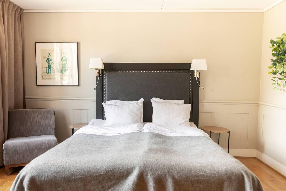 Ein Hotelzimmer mit einem großen Bett, zwei Nachttischlampen, einem gerahmten Bild an der Wand, einem grauen Sessel und einem kleinen Tisch.