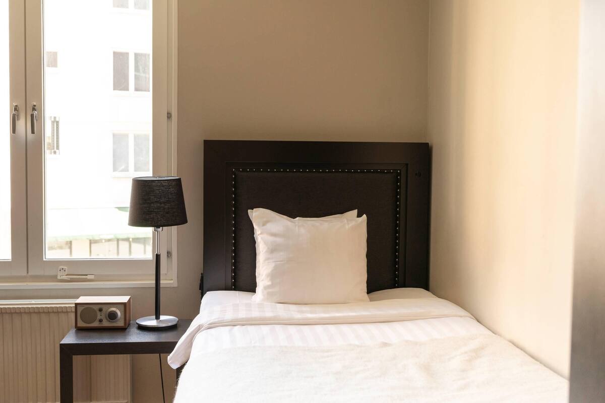 Ein ordentlich bezogenes Bett mit dunklem Kopfteil, ein Nachttisch mit Lampe und Radio sowie ein Fenster mit Jalousien.