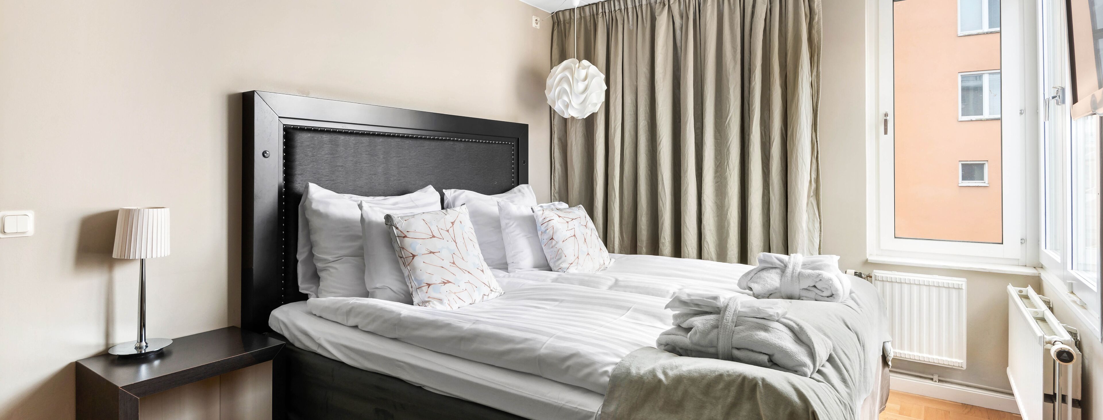 A neatly made bed with white linens and decorative pillows, a bedside table with a lamp, and a view of a building through a window with beige curtains.