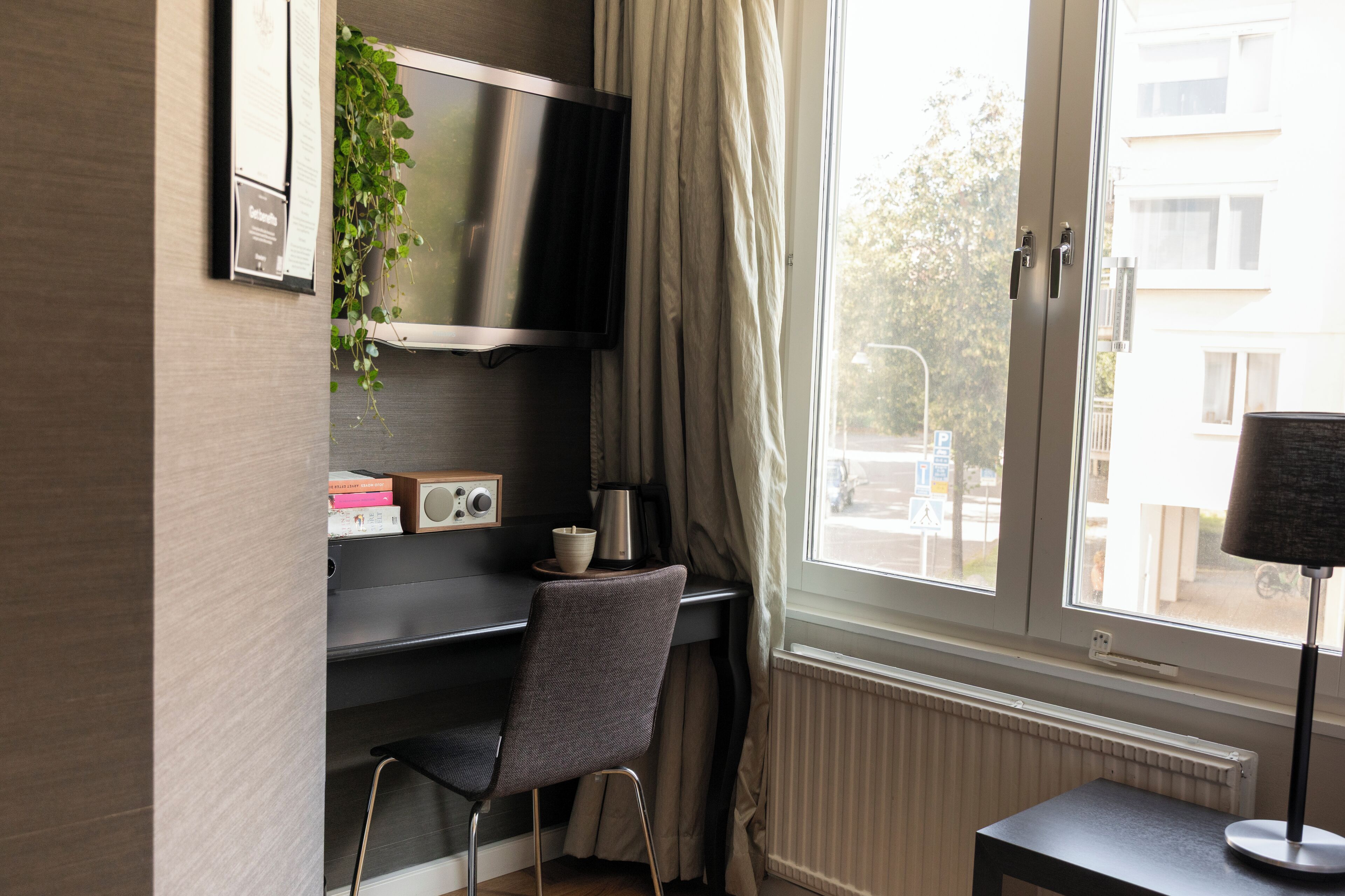Una habitación con escritorio, silla, televisión, ventana con cortinas y radiador.