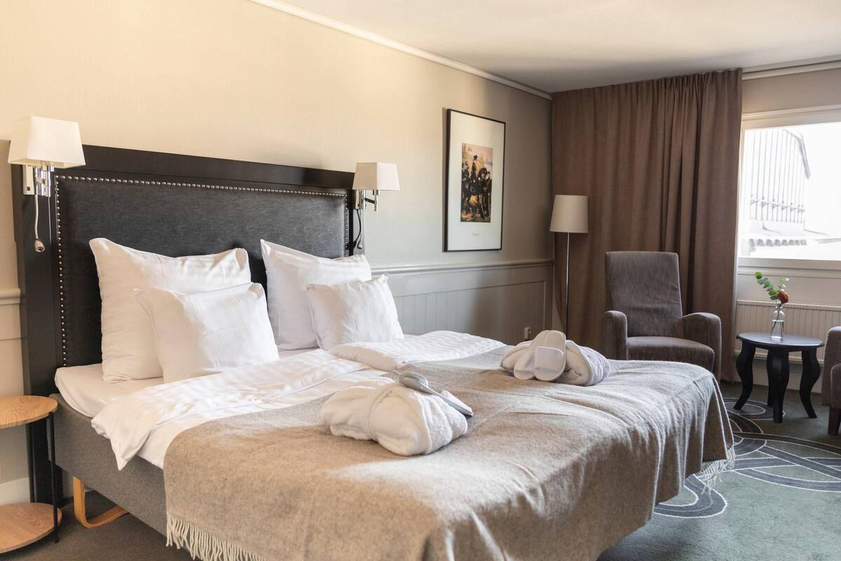 Ein Hotelzimmer mit einem großen Bett, einem Sessel, einem kleinen Tisch und einem Fenster mit Vorhängen.